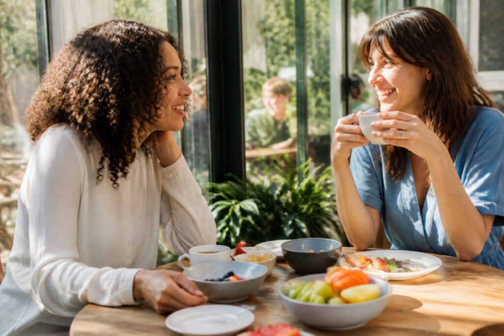 Duas amigas conversando e rindo num café da manhã ensolarado, com xícaras e frutas na mesa