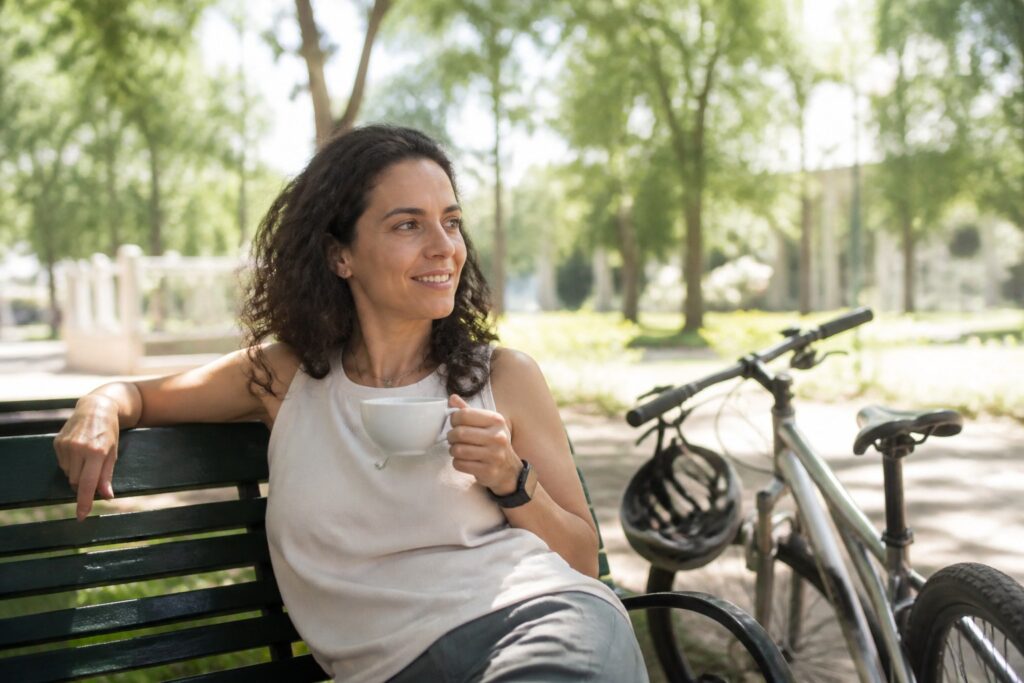 Mulher descansada e sorridente tomando café num banco de praça numa manhã de domingo ensolarada, com a bicicleta ao lado