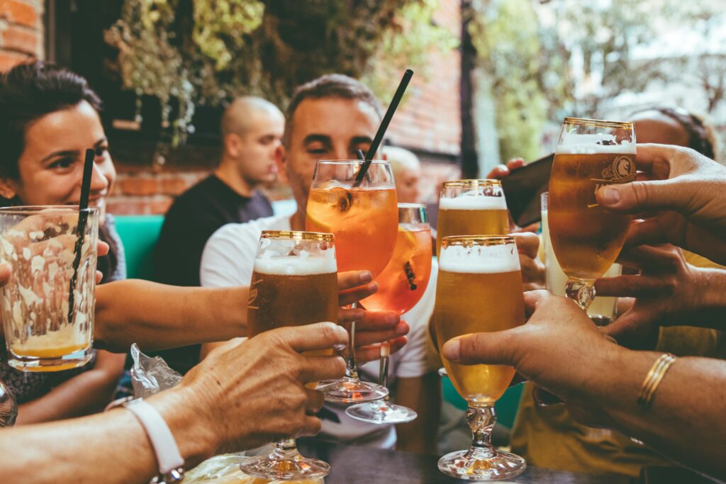Mãos de várias pessoas erguendo copos de cerveja e drinks num brinde, num bar